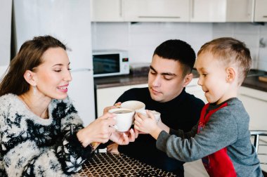 Genç anne, baba ve çocuk sabahları kahve ve çay içiyor. Mutlu anne, baba ve oğul evdeki mutfakta kahvaltı yapıyorlar. Aile, yemek ve insan konsepti.