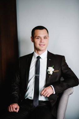 Gelinliğinde yaka çiçeğiyle beyaz arka planda, koltukta oturan çekici, mutlu damat. Erkekler kameraya bakıyor..