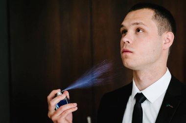 Damat şişesinin parfüm sıkması, gürültü. Zarif, genç, çekici, şık, stil sahibi, takım elbiseli, kahverengi arka planda duran parfüm ya da parfüm kullanan bir adam..