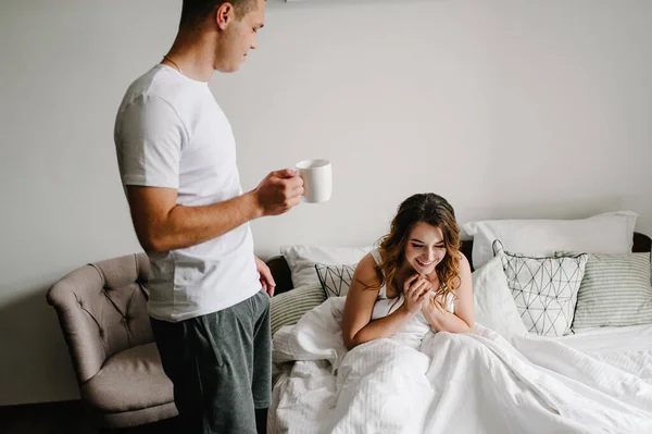 Loving husband carries coffee for his happy wife in the bed, in a big bedroom, side view. The Valentine\'s Day Concept. Celebrating holiday. Lifestyle. Women\'s Day, eighth of March.