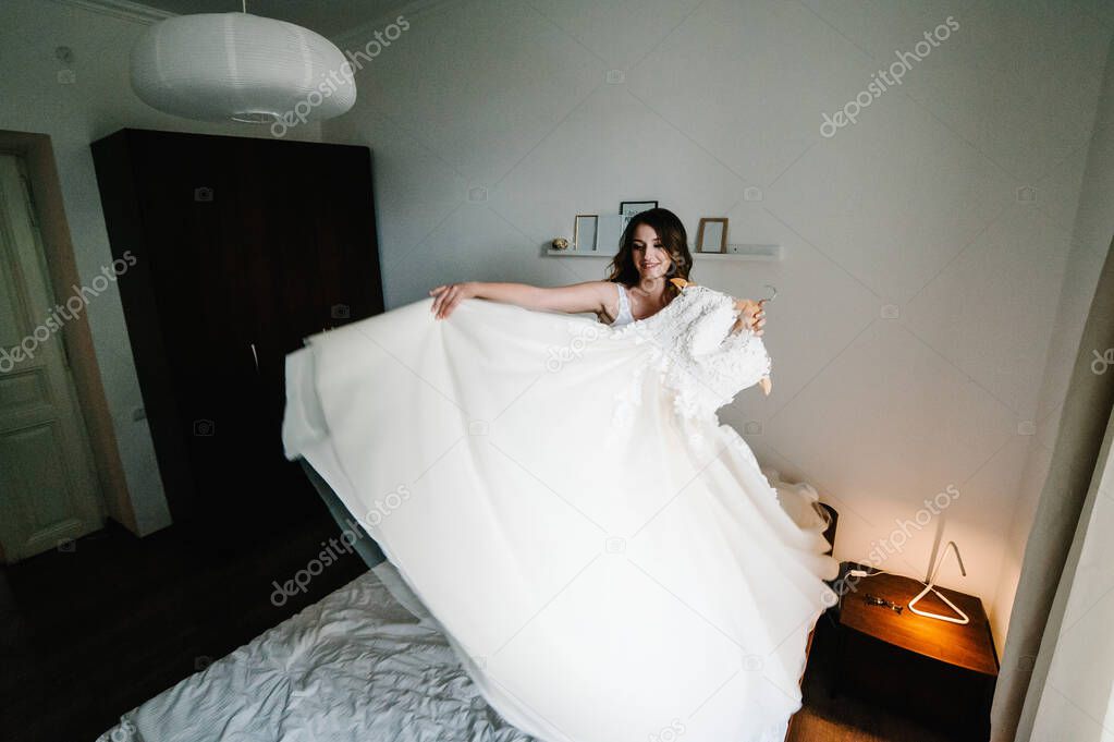 The bride holds a wedding dress in her hands and spins, turn with her on the bed. Style vintage elegant dress with lace. Wedding Morning Preparations.