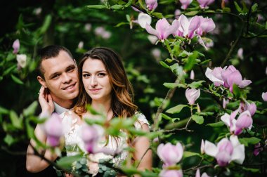 Pembe ve mor manolya ve yeşillik çiçeklerinin arka planında parkta kucaklaşan ihtiraslı yeni evliler. Düğünün yapılacağı yer..