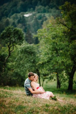 Bekle bebeğim. Göbeğe bakıyorum. Sevgili kocası olan hamile bir kadın ağacın yanındaki çimlerin üzerinde oturuyor. Yuvarlak göbek Samimi ve hassas an. Arka plan, doğa, park, ağaç, orman, dokuz ay. mutluluk