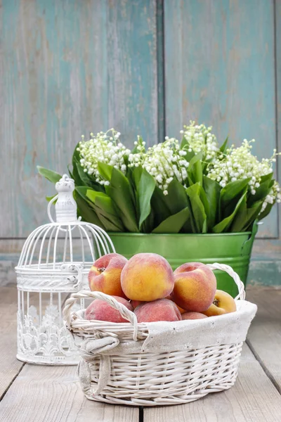 Basket of peaches on wooden table, bucket with lily of the valle — Stock Photo, Image