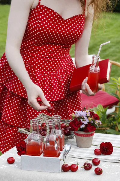 Femme en robe rouge tenant une cerise mûre, bouteille de jus de cerise — Photo