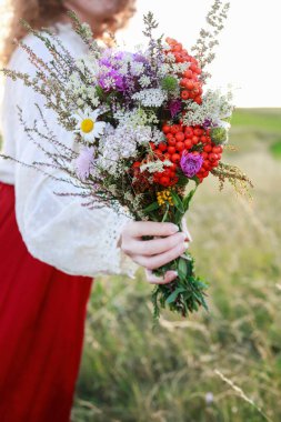 Kırmızı etekli genç bir kadın çayırdan harika bir buket yabani çiçek tutuyor. Parti dekoru