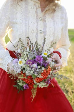 Kırmızı etekli genç bir kadın çayırdan harika bir buket yabani çiçek tutuyor. Parti dekoru