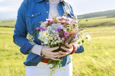 Kot ceketli genç kadın elinde çayırdan büyük, çarpıcı bir buket kır çiçeği tutuyor. Parti dekoru