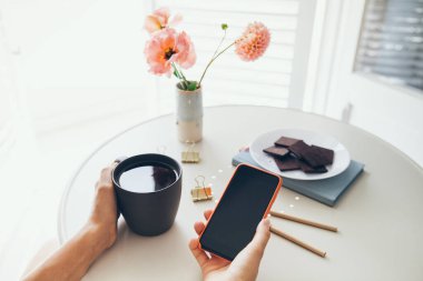 Close up of woman hands holding cup of tea and mobile phone, reading news, social network, surfing the web in while staying at home. Remote work, video confidence calls with colleagues.