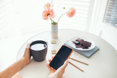 Akıllı telefon tutan, haber okuyan, sosyal ağlar kuran, çay içerken görüntülü konuşma yapan ve çikolata yiyen kadınların ellerini kapatın. Not defteri, kalemler ve vazo dolusu çiçekler masanın üstünde..