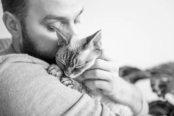 Siyah beyaz sakallı genç adamın sevimli Devon Rex kedisiyle sarılıp yattığı fotoğraf. Kedilerle vakit geçirmek, moralini yükseltmek ve stres seviyeni düşürmek. Sevgiyi bir evcil hayvanla paylaşmak