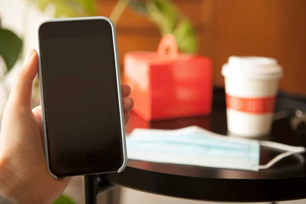 Close-up photo of woman hand holding smartphone, using food delivery service application while staying home at quarantine. Checked out the restaurants. Home interior background with takeaway food.
