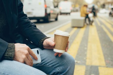 Şehrin sokaklarındaki bankta oturan ve kahve molası veren erkek ellerine yakın çekim. El ele tutuşmak bir fincan kahveyi alıp götürür, rahatlatır, çevrimdışı yapar. Seçici odak