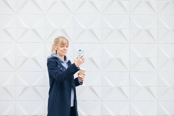 Woman is standing on concrete modern architecture wall background dressed in casual clothes blue color hoodie and coat holding smart phone in hand and having video call.