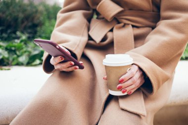 Close up of woman hands holding mobile phone, using internet while sitting outdoors on the bench and drinking speciality coffee. Chatting with friends and family, reading latest news and updates in the web.