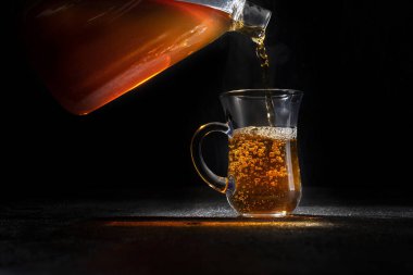 Siyah arka planda şeffaf bir fincana sıcak çay dökülüyor..