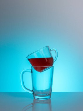 Kırmızı bir sıvıyla dolu temiz bir cam bardak, başka bir boş fincana monte edilmiş bir masa üzerinde bir mat bitiş gün ışığı ışınları, yakın plan, renkli bir arka plan.