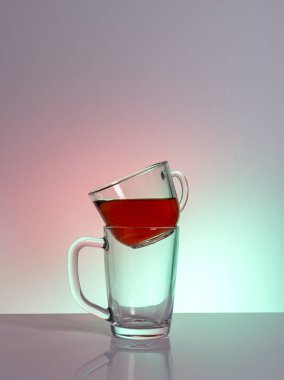 Kırmızı bir sıvıyla dolu temiz bir cam bardak, başka bir boş fincana monte edilmiş bir masa üzerinde bir mat bitiş gün ışığı ışınları, yakın plan, renkli bir arka plan.