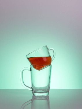 Kırmızı bir sıvıyla dolu temiz bir cam bardak, başka bir boş fincana monte edilmiş bir masa üzerinde bir mat bitiş gün ışığı ışınları, yakın plan, renkli bir arka plan.