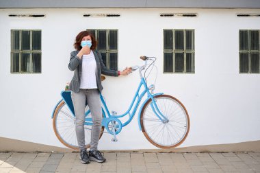middle-aged woman with face mask leaning against a wall with painted bicycle