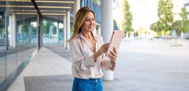 Professional woman scrolling on tablet while walking through modern city street