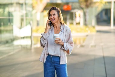 Businesswoman walking in urban setting, talking on mobile phone and holding coffee cup