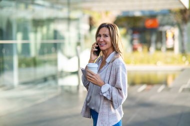 Woman standing outdoors smiling and talking on smartphone while holding a coffee cup
