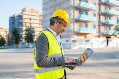 Engineer man wearing hard hat and safety vest, working on laptop at building site