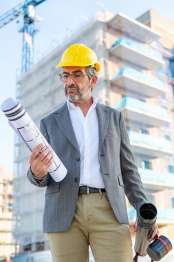 Mimarın elinde bina planı var. İnşaat projesini kontrol ediyor.