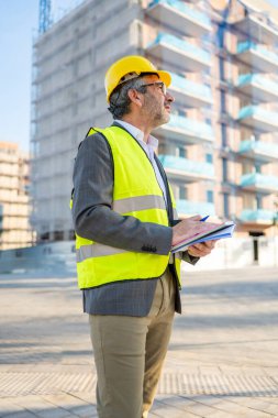 İnşaat yöneticisi elinde pano bulunan inşaat alanını inceliyor