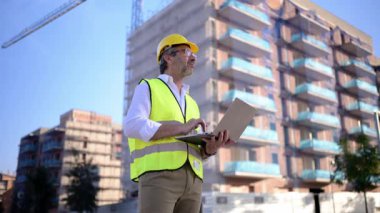 Kasketli ve güvenlik yeleği giymiş bir erkek mimar inşaat alanında dizüstü bilgisayarla çalışıyor.