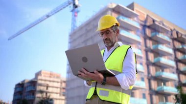 Kasketli ve güvenlik yeleği giymiş bir erkek mimar inşaat alanında dizüstü bilgisayarla çalışıyor.