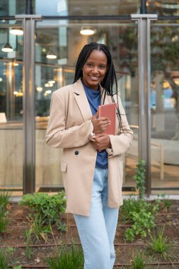Black woman smiling, holding a book, standing in front of a modern building