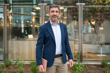 Olgun bir iş adamı, elinde tabletle kurumsal bir cam binanın önünde dururken gülümsüyor.