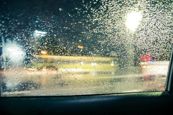 Road view through car window with rain drops at night. Defocussed traffic viewed through a car windscreen covered in rain, focus on raindrops