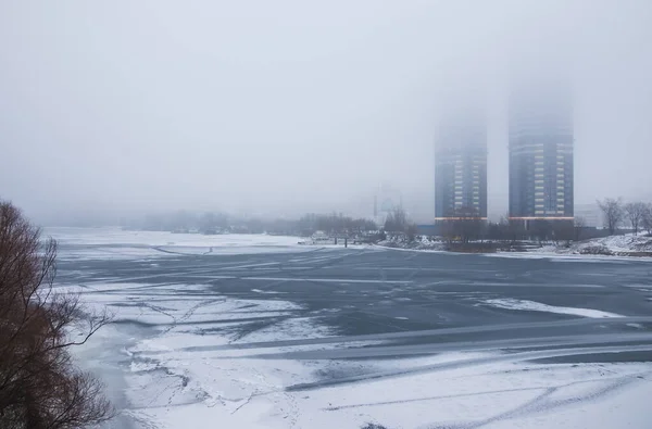 Winter landscape. Frozen river in fog on a cloudy day. Tall buildings in the fog on the river bank. Dnipro. Kyiv, Ukraine