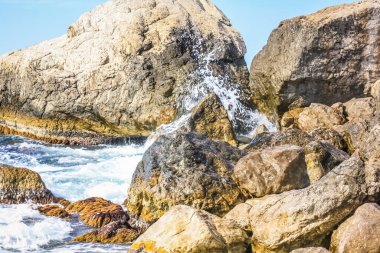 Turkuaz deniz taşı plajı, parlak güneş ışığında dalgalar kırılıyor. Renkli parlak deniz arka planı. Yaz konsepti, tatil, seyahat. Saf deniz suyu, sahilde büyük taşlar.