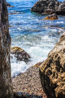 Turkuaz deniz taşı plajı, parlak güneş ışığında dalgalar kırılıyor. Renkli parlak deniz arka planı. Yaz konsepti, tatil, seyahat. Saf deniz suyu, sahilde büyük taşlar.