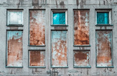 Pencereler yerine paslı plakaları olan eski gri bir binanın cephesi