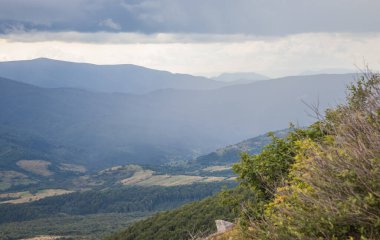 Katmanlı dağ vadilerine bakan kayalık bir uçurum. Huysuz gökyüzü ve uzak tepeler sise dönüşür, desenli manzaralar ve alp çimenlerinin çerçevesi tamamen düşer, hisseder