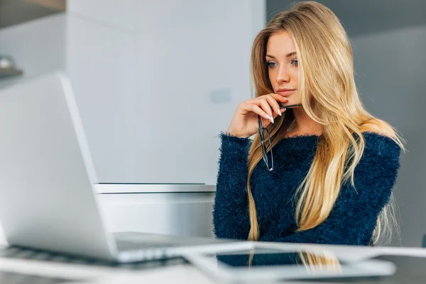 Ung kvinde sidder i køkkenet med laptop. Hun holder e - Stock-foto