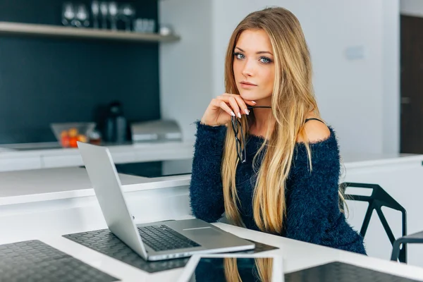 Smuk ung kvinde sidder i køkkenet med laptop. Det er hun. - Stock-foto