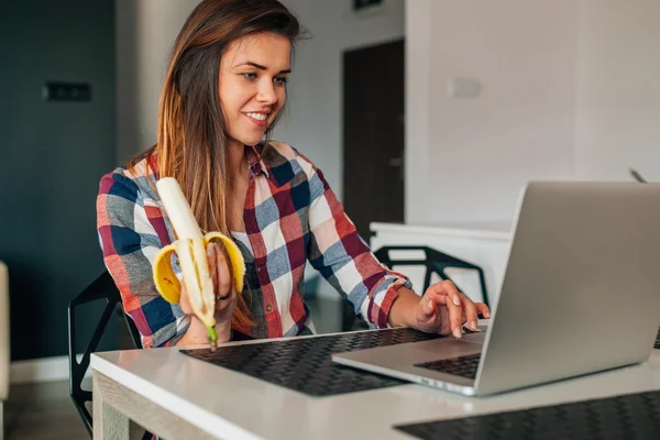 Dziewczyna siedzi i kuchni i za pomocą laptopa. Ona jest jedzenie banan — Zdjęcie stockowe