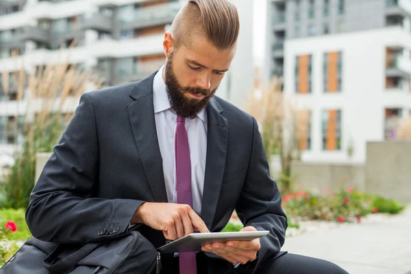 Jonge, bebaarde zakenman zittend op een bankje en de tablet pc gebruiken — Stockfoto