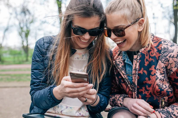 Dvě dívky sedí v parku a pomocí mobilního telefonu. — Stock fotografie
