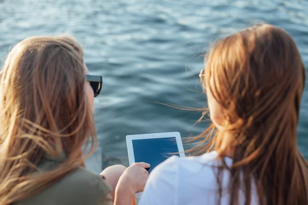两个年轻女孩坐在旁边的河和使用平板电脑. — 图库照片