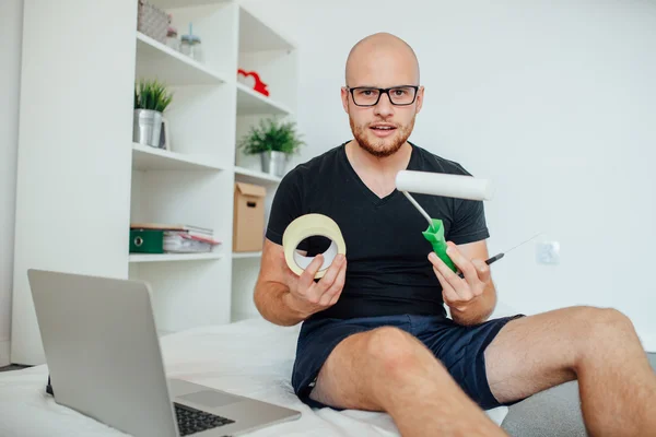 Young man is sitting on the bed and holding painting tools. Home — Stock Photo, Image