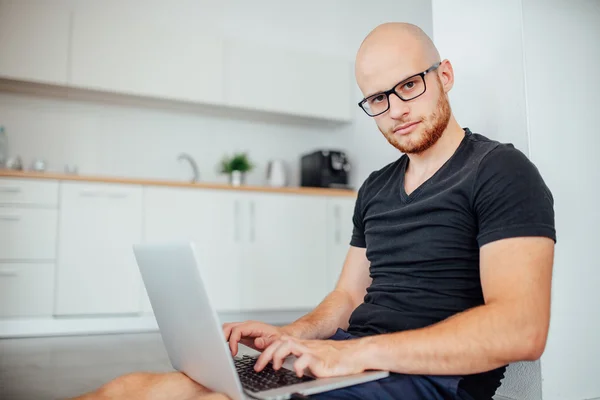 Młody człowiek jest za pomocą jego laptopa pc i siedzi w kuchni. — Zdjęcie stockowe