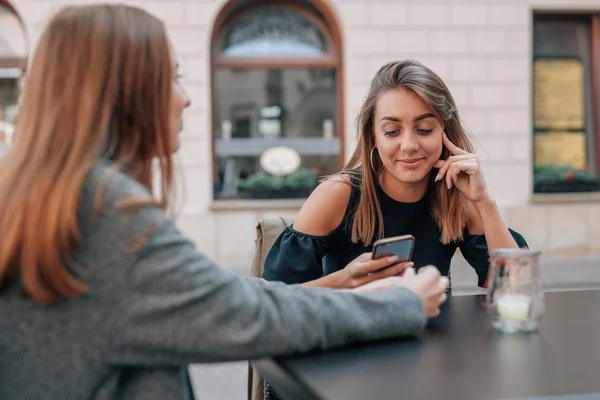 Unga kvinnor sitter på café och med hjälp av mobiltelefon. Staden Stockfoto