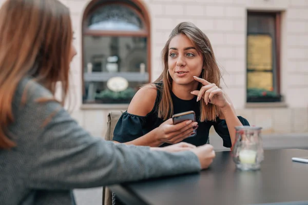 Unga kvinnor sitter och använder mobiltelefon. Utomhus café bac Stockbild